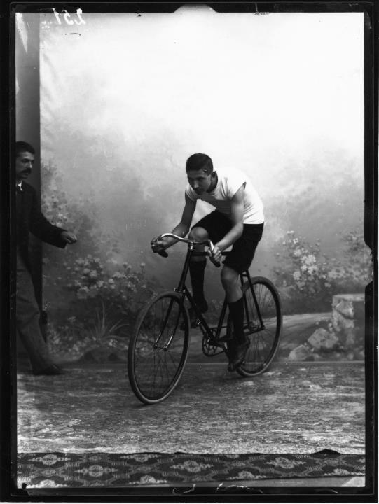 Ripresa in studio. Ritratto del sig. Friedrich Held sen. (nato 1876 a Bolzano, morto 1963 a Innsbruck, commerciante) su una bicicletta. Al lato si vede un collaboratore dello studio fotografico Waldmüller, che sta stabilizzando il ciclista con un filo metallico