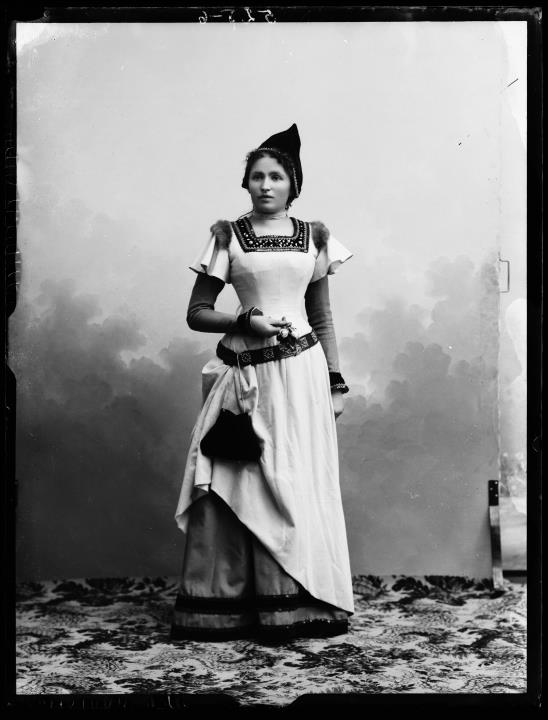 Ripresa in studio. Ritratto di una donna con vestito in piedi con cintura e borsa. Tiene dei fiori in mano.