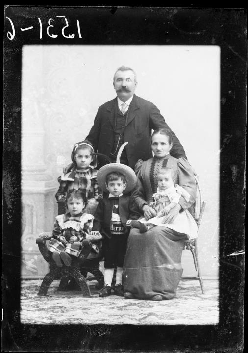 Studioaufnahme. Gruppenporträt eines Mannes, einer Frau und vier Kindern. Der Bub in der Mitte trägt Lederhose und einen Gurt mit der Aufschrift 