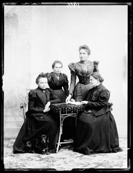 Studioaufnahme. Gruppenporträt, vier Frauen, die an einem Tisch sitzen und stehen. Auf dem Tisch ist ein offenes Buch