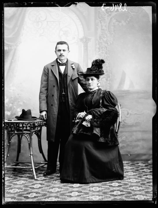 Studioaufnahme. Gruppenporträt einer Frau und eines Mannes mit Hut und Pelz. Auf dem Boden ein Teppich, darauf ein Tisch