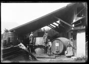 Ripresa in esterno. Tre uomini davanti a botti di legno in un bottaio. Su una botte l'iscrizione 