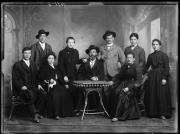 Ripresa in studio. Ritratto di gruppo di quattro donne e cinque uomini, quattro persone sono sedute, in primo piano un tavolo. Tre uomini indossano un cappello