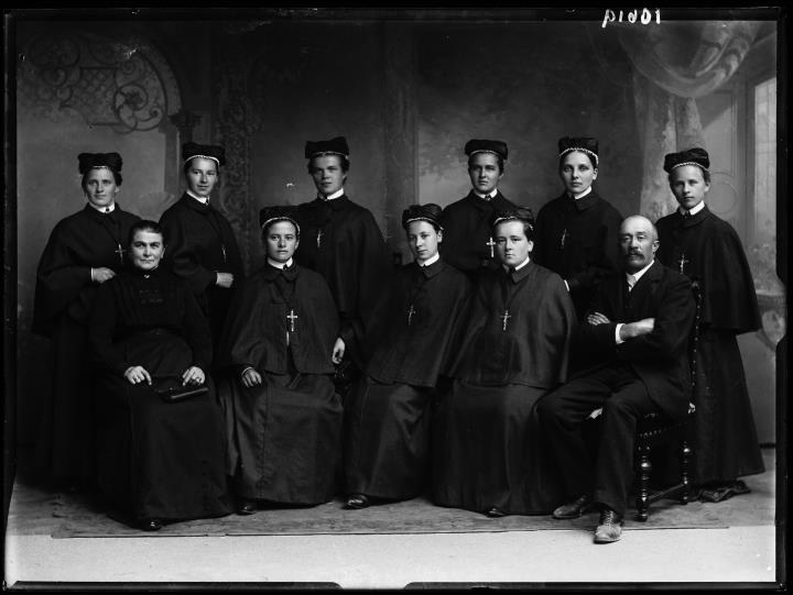 Studioaufnahme Gruppenporträt, zehn Frauen, ein Mann. Neun Frauen tragen Ketten mit Kreuz und Hüte