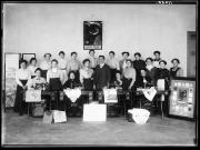 Ripresa in interno. Ritratto di gruppo di un uomo e 20 donne. In primo piano ci sono quattro macchine da cucire e uno scudo con la scritta 