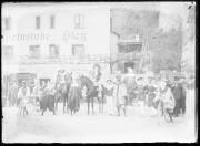 Ripresa in esterno. Ritratto di gruppo, gruppo di uomini, donne e bambini, due cavalli, tre biciclette. Alcune persone portano un costume. Sullo sfondo un palazzo con la scritta 