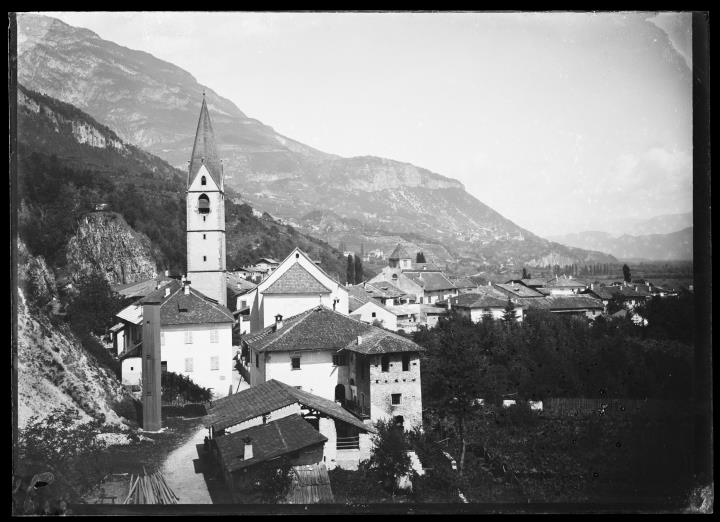Außenaufnahme. Das Dorf Margreid: Mehrere Gebäude, eine Kirche, ein Taubenschlag und ein Obstgarten. Im Hintergrund ein zweites Dorf und Berge.
