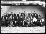 Foto di gruppo di 43 uomini e un ecclesiastico al centro. Sopra, le tende che regolano la luce naturale nello studio fotografico