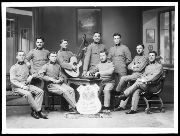 Studioaufnahme. Gruppenporträt von neun Soldaten in Uniform, davon einer mit Gitarre mit zwei Hälsen. In der Mitte eine Tafel mit 