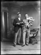 Ripresa in studio. Ritratto di gruppo di due uomini in piedi con bicchieri di birra in mano. Uno die due porta un´uniforme, un cappello e un mazzo di fiori. L´altro porta un berretto e un fazzoletto da collo con la scritta 