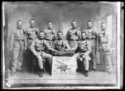 Riproduzione. Ripresa in studio. Ritratto di gruppo: nove soldati, uno di loro con una tromba. In mezzo una lavagna con la scritta 