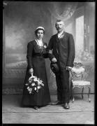 Ripresa in studio. Ritratto di gruppo: un uomo e una donna. Lei tiene in mano un mazzo di fiori e indossa una coroncina bianca nei capelli. L'uomo tiene in mano un cappello. Entrambi hanno dei fiori attaccati sui vestiti