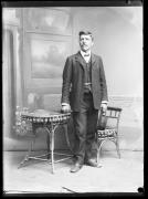 Ripresa in studio. Ritratto di un uomo in piedi con un cappello in mano. Vicino ci sono un tavolo e una sedia