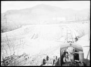 Ripresa in esterno. Cantiere, macchina sui binari. Sullo sfondo un pilone dell'acqua, case, cavi dell'elettricità, un stanga chiusa e un casellante