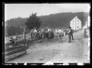 Außenaufnahme. Straße, Männer und Frauen stehen an einem Brunnen. In Hintergrund ein Haus, eine Kirche