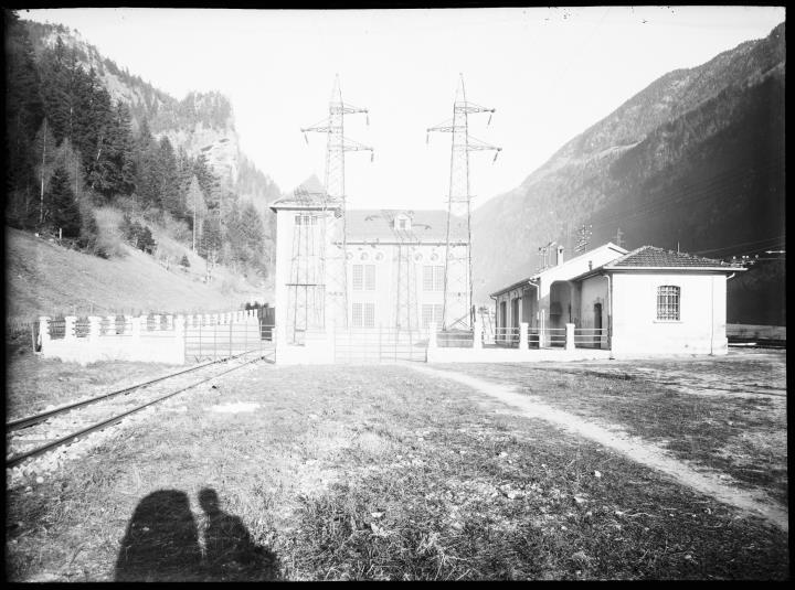Außenaufnahme eines Elektrizitätswerkes. Im Vordergrund links Gleise. Zwei Strommasten. Vorne der Schatten des Fotografen mit seiner Kamera