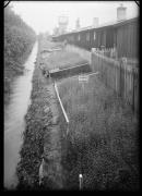 Ripresa in esterno. Giardino con ginestra, canale e casetti di legno. Insegne con le iscritte 