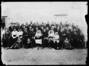 Ripresa in esterno. Ritrato di gruppo, festeggiamento per l'ordinazione sacerdotale, uomini, donne e bambini. Una casa con l'iscrizione 