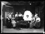 Ripresa in studio. Ritratto di gruppo di 13 donne e un uomo. Nella parte superiore i tendaggi con cui regolare l'incidenza della luce. Dietro al gruppo sono montato due fondali dipinti. La luce bianca attorno alla donna seduta al tavolo è frutto di un ritocco; Sul negativo la figura era stata scontornata con un colore scuro coprente, che nell'inversione del positivo è diventato bianco. Cosí isolato, il ritratto poteva essere utilizzato anche separatamente, magarin per un cameo o un annuncio funebre