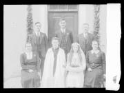 Ripresa in esterno. Ritratto di gruppo di cinque uomini e due donne davanti ad una porta. Una donna tiene un cuscino con un calice sopra